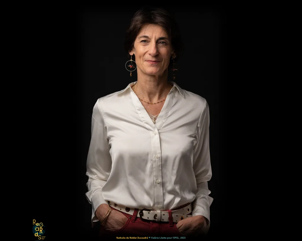 Portrait de Nathalie de Noblet-Ducoudré debout en chemisier de satin blanc, mains dans les poches, sur fond noir.