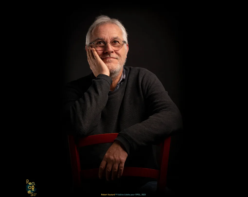 Portrait de Robert Vautard, cheveux blancs et lunettes, la joue dans la main, assis sur une chaise rouge, le regard levé.