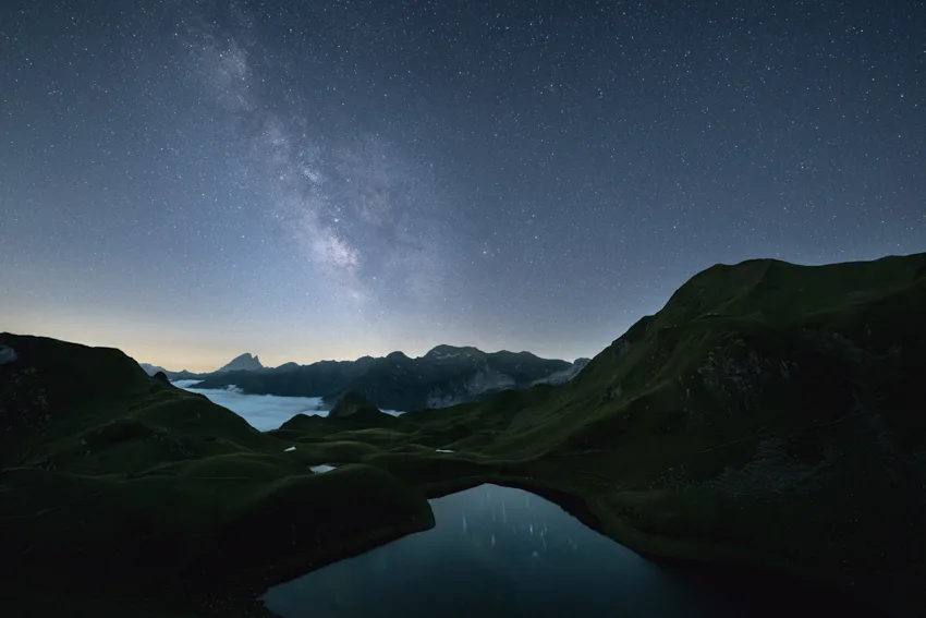 Nuit étoilée sur le lac de Montagnon : la Voie lactée au-dessus des crêtes, une mer de nuages au loin et le lac au premier plan.
