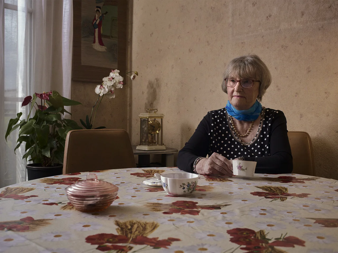 Une femme âgée, seule à une table couverte d'une nappe fleurie, tient une tasse de thé dans un intérieur aux murs beiges.