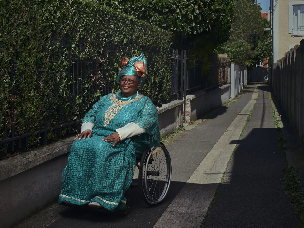 Une femme en fauteuil roulant, en robe et turban turquoise, sourit sur un trottoir étroit bordé d'une haie.
