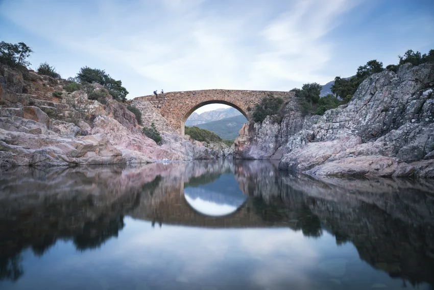 Pont de pierre à une arche au-dessus d'une rivière de Corse aux eaux calmes, entre des rochers clairs, l'arche se reflétant dans l'eau.