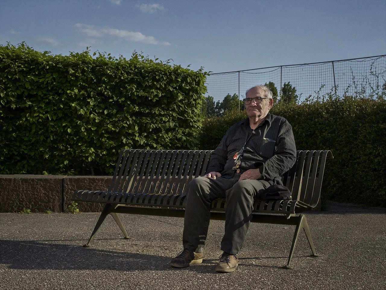 Un homme âgé à lunettes, en chemise sombre, assis seul sur un banc devant une haie, dans la lumière dorée du soir.