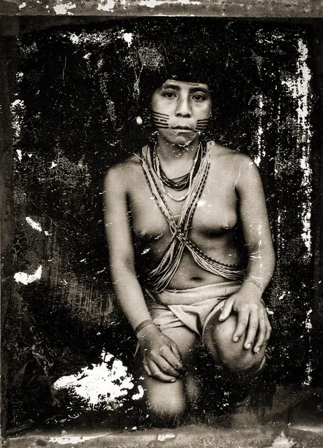 Portrait sépia d'une jeune femme d'Amazonie agenouillée, torse nu, colliers croisés sur la poitrine et peintures rayées sur le visage.
