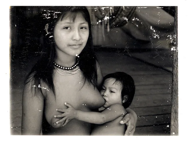 Portrait sépia d'une jeune mère d'Amazonie, collier de perles au cou, allaitant un enfant qu'elle tient contre elle.