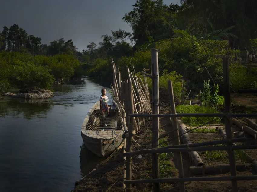Une enfant debout dans une longue barque en bois amarrée au bord d'un canal, le long d'une clôture de bambou, à la tombée du jour.
