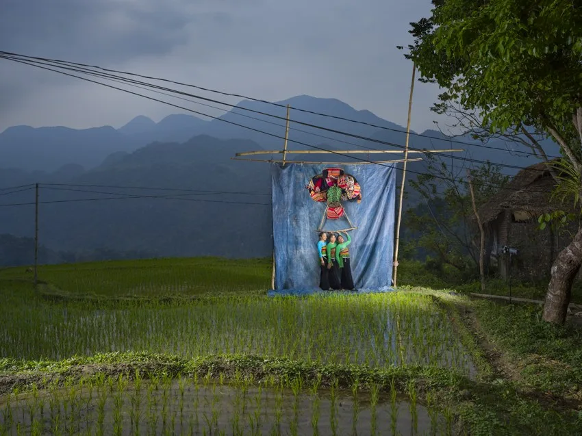 Trois femmes en tenue traditionnelle posent devant une bâche bleue ornée de textiles colorés, au milieu des rizières et des montagnes.
