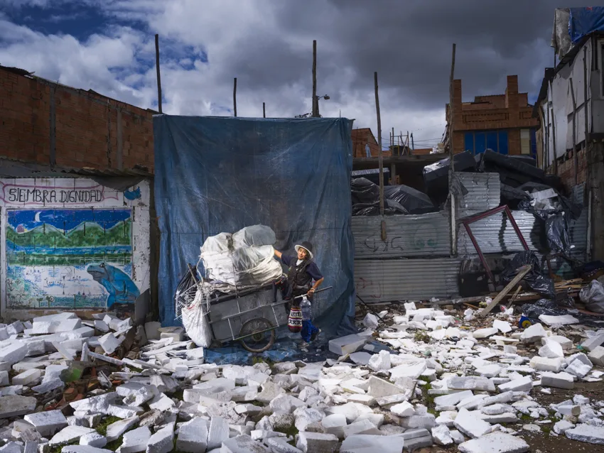Une femme en chapeau pousse une charrette chargée de sacs blancs au milieu de débris blancs, devant une bâche bleue et une fresque.