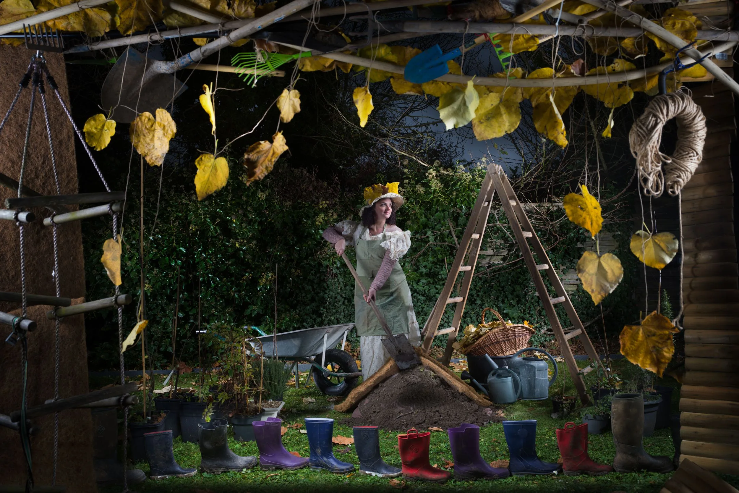 Une femme en robe et chapeau jaune, pelle en main, dans un décor de jardin avec échelle, outils, feuilles suspendues et bottes alignées.
