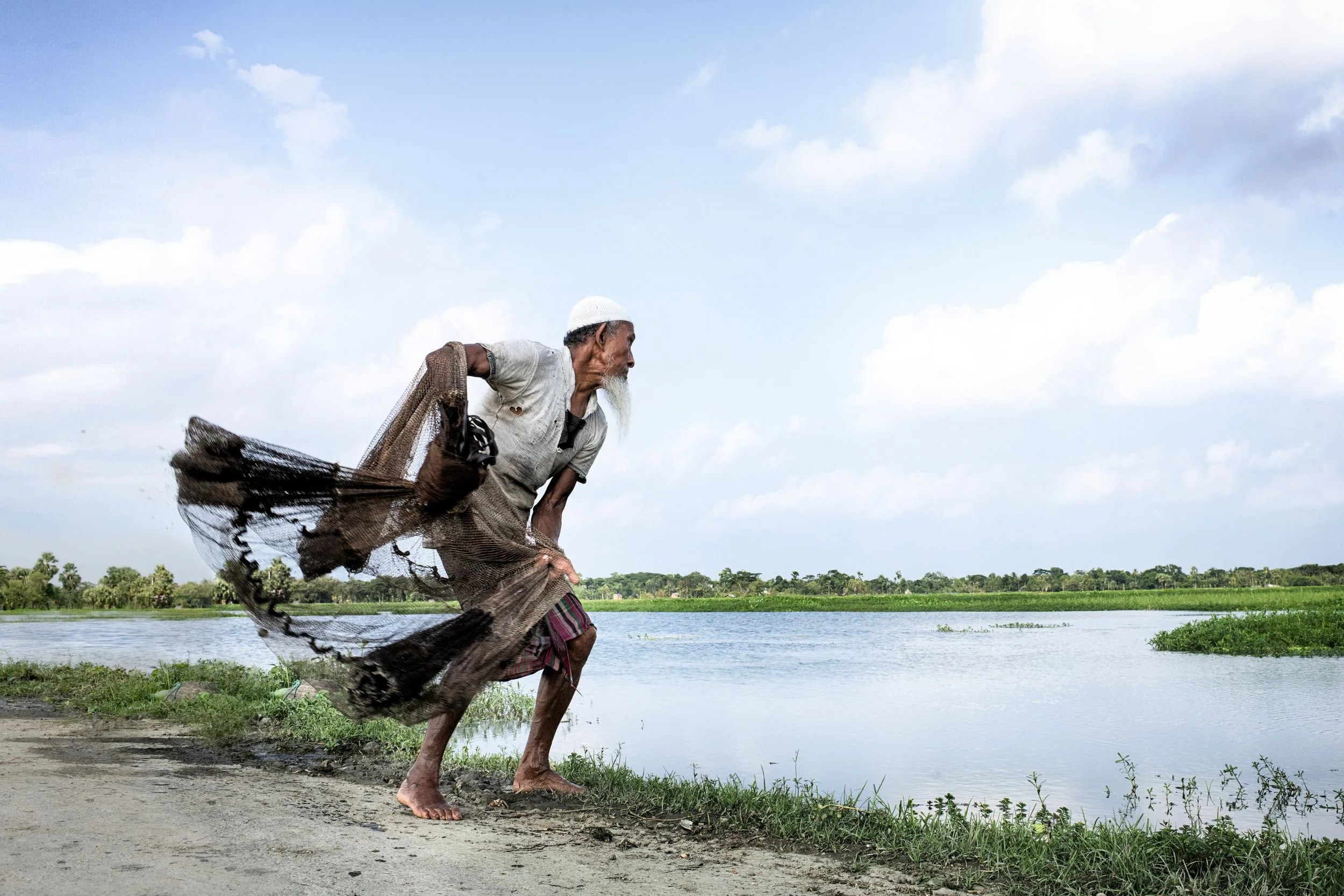 Un homme à barbe blanche et calotte blanche, pieds nus, lance un filet de pêche au bord d'une étendue d'eau, sous un ciel nuageux.