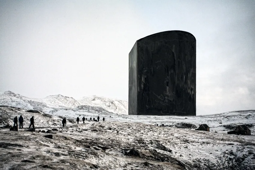 Un immense monolithe noir posé sur un paysage arctique enneigé, une file de marcheurs avance à ses pieds.
