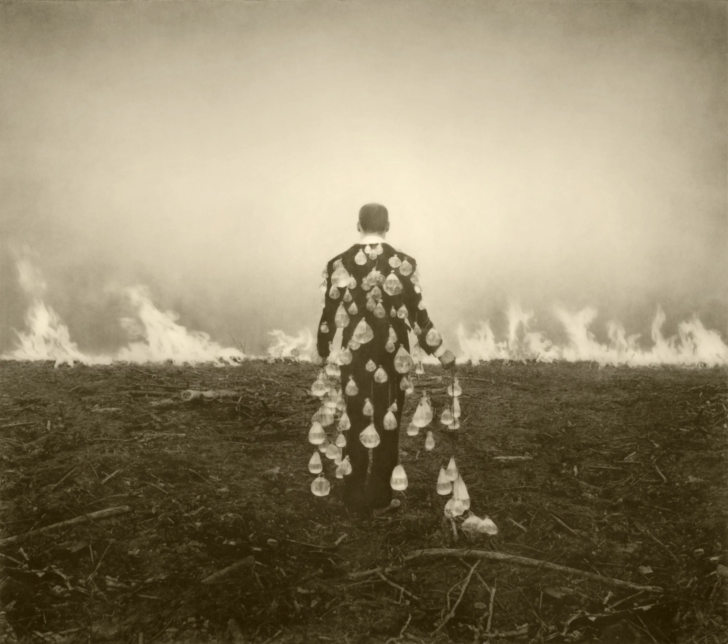 Un homme en costume, de dos, couvert de gouttes de verre, marche vers un champ en flammes, œuvre de Robert et Shana ParkeHarrison.