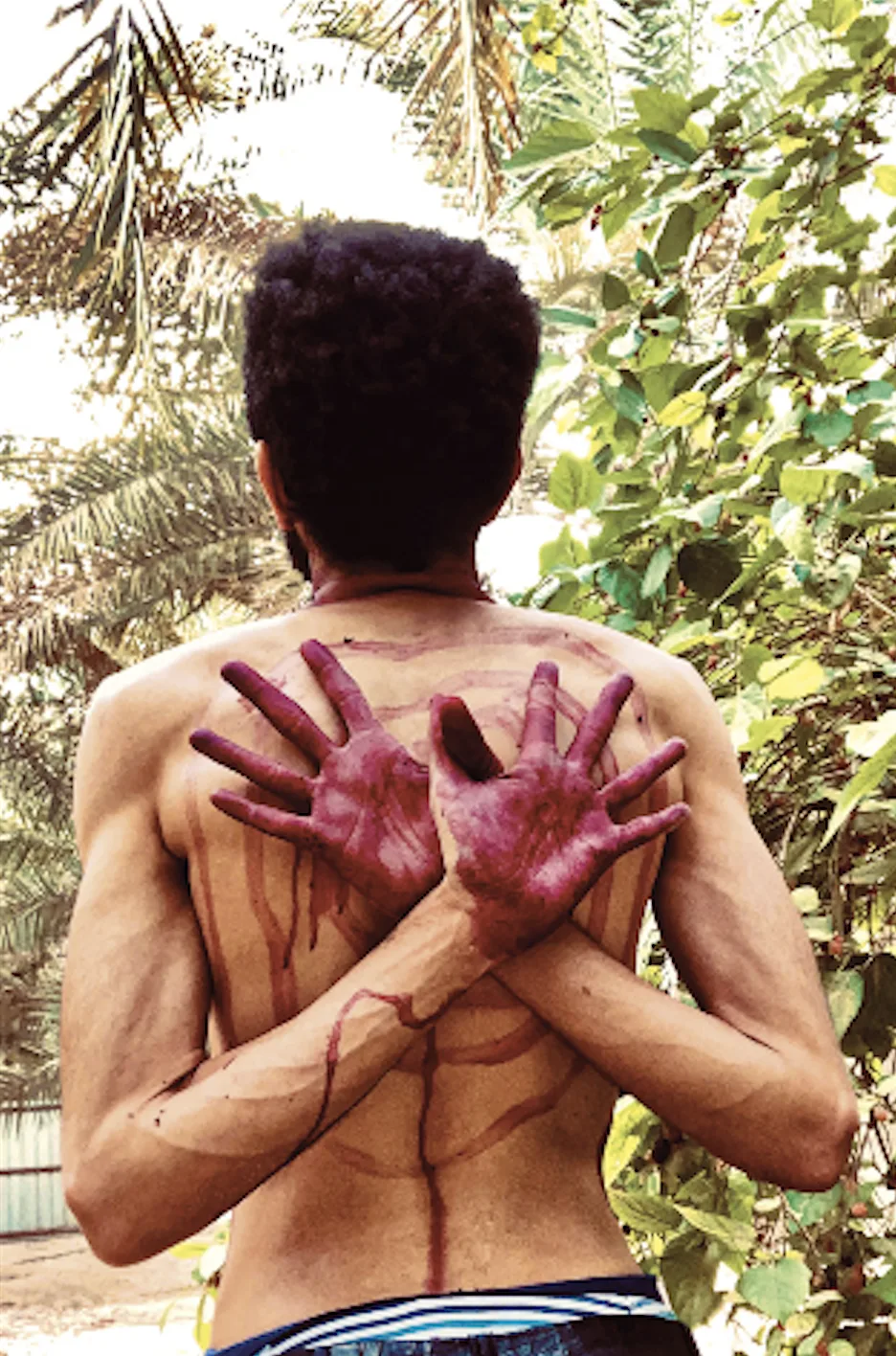 Un homme torse nu, de dos, les bras croisés et les mains teintes de rouge posées sur ses épaules, devant des palmiers, par Mohamed Alfaraj.