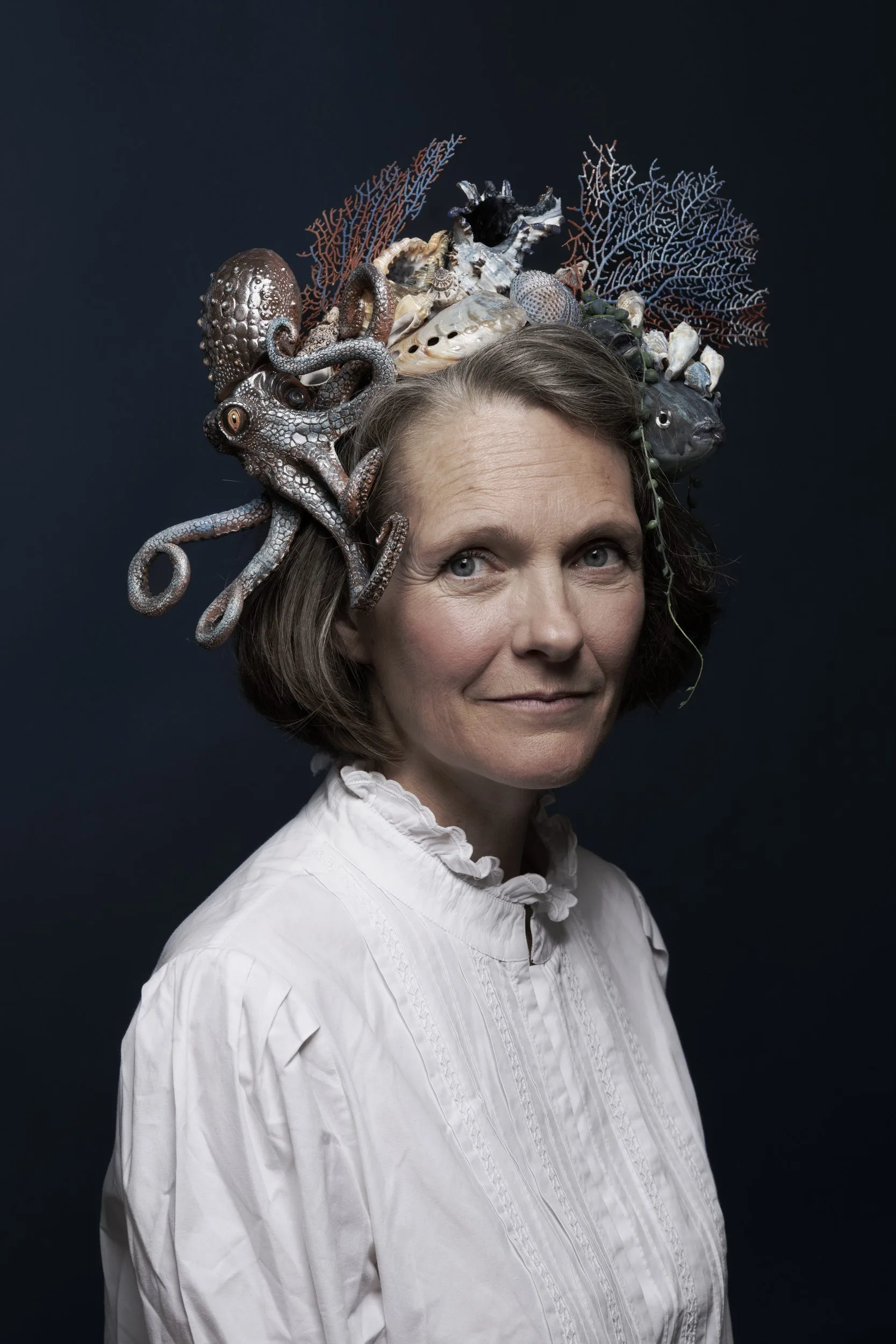 Portrait de Claire Nouvian en chemisier blanc, coiffée d'une couronne de corail, de coquillages, d'un poulpe et d'un poisson, sur fond bleu nuit.
