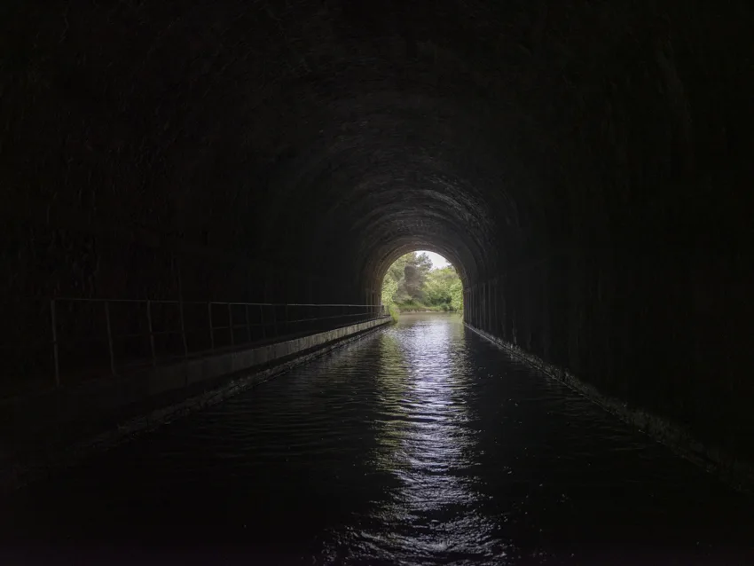 L'intérieur sombre d'un tunnel de canal voûté, avec son chemin de halage, l'eau menant vers une ouverture verte et lumineuse.