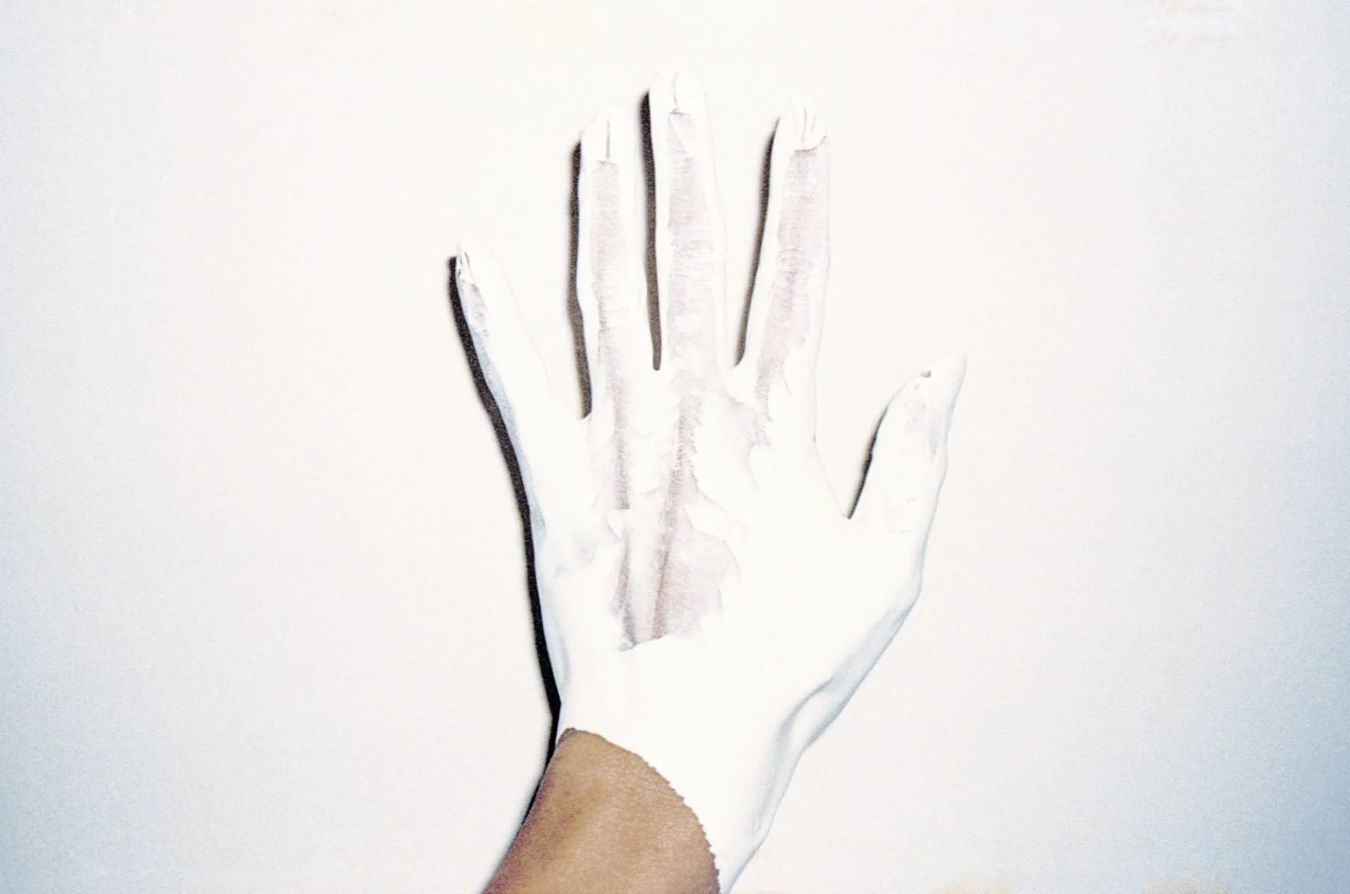 Une main couverte de peinture blanche, doigts écartés, posée contre un mur blanc, seul le poignet garde sa couleur.