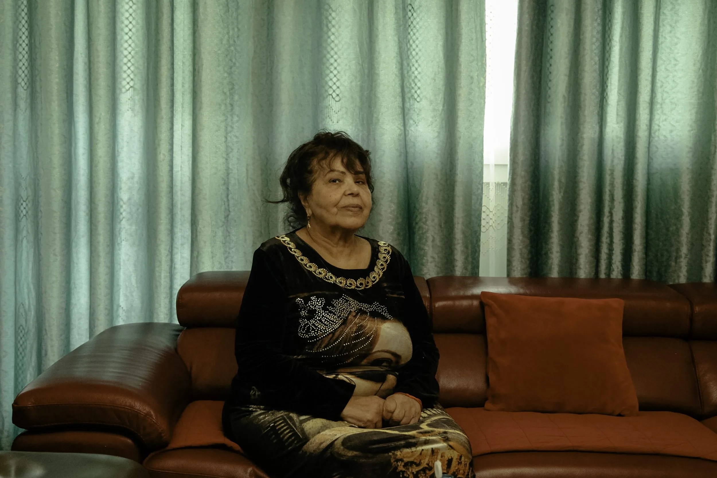 Portrait d'une femme assise sur un canapé en cuir brun, en haut noir imprimé d'un visage, devant de grands rideaux verts, par Lydia Saidi.