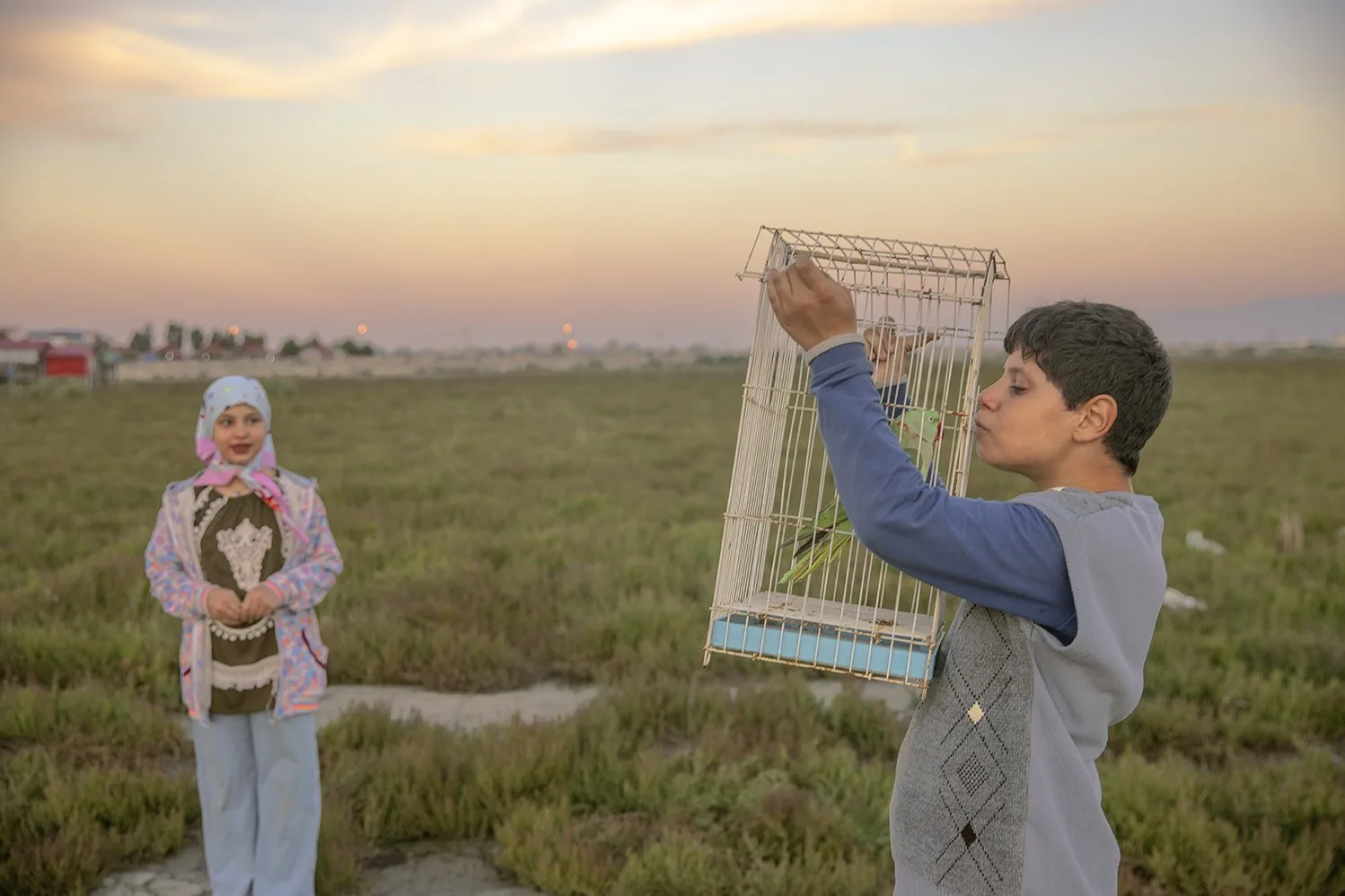 Au coucher du soleil, un garçon lève une cage à oiseaux dans un champ, une fillette au foulard derrière lui, par Masoumeh Bahrami.