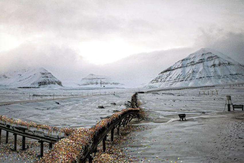 Paysage arctique enneigé où une conduite sur pilotis disparaît sous des confettis multicolores, des montagnes au fond.