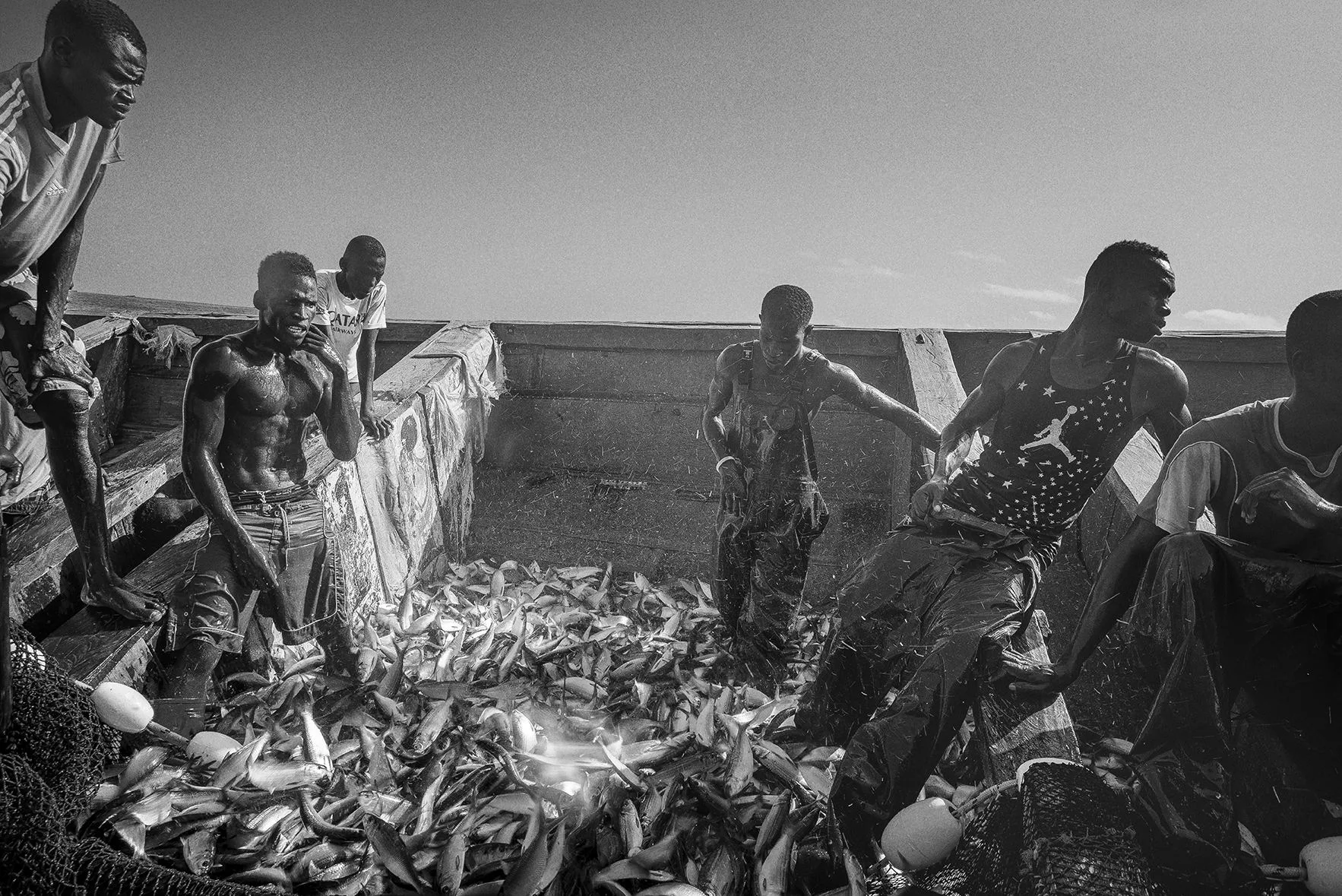 Noir et blanc : des pêcheurs debout dans une grande pirogue remplie de poissons argentés qui brillent dans la lumière.