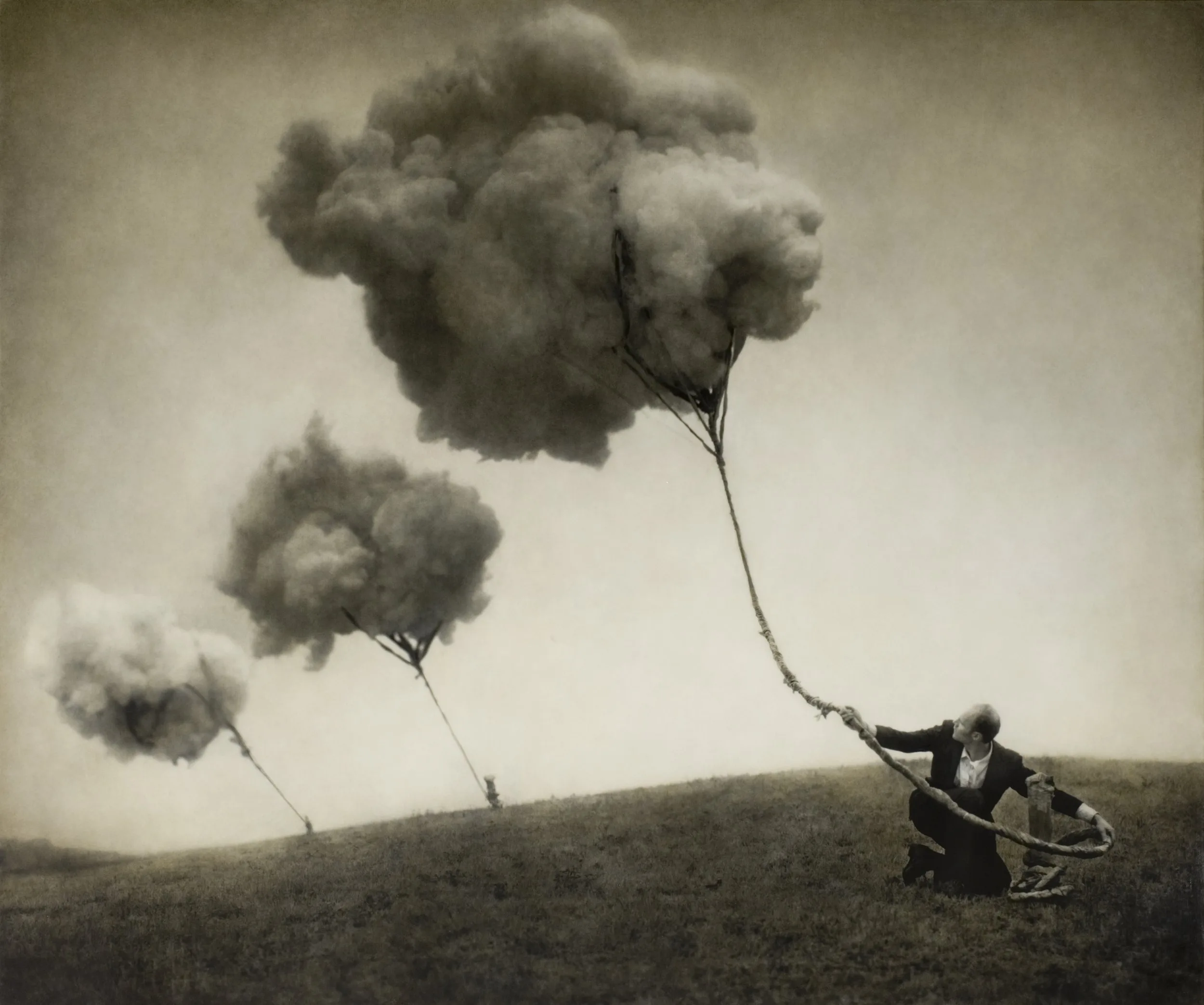 Un homme en costume agenouillé sur une colline retient par des cordes trois nuages qui flottent dans le ciel.