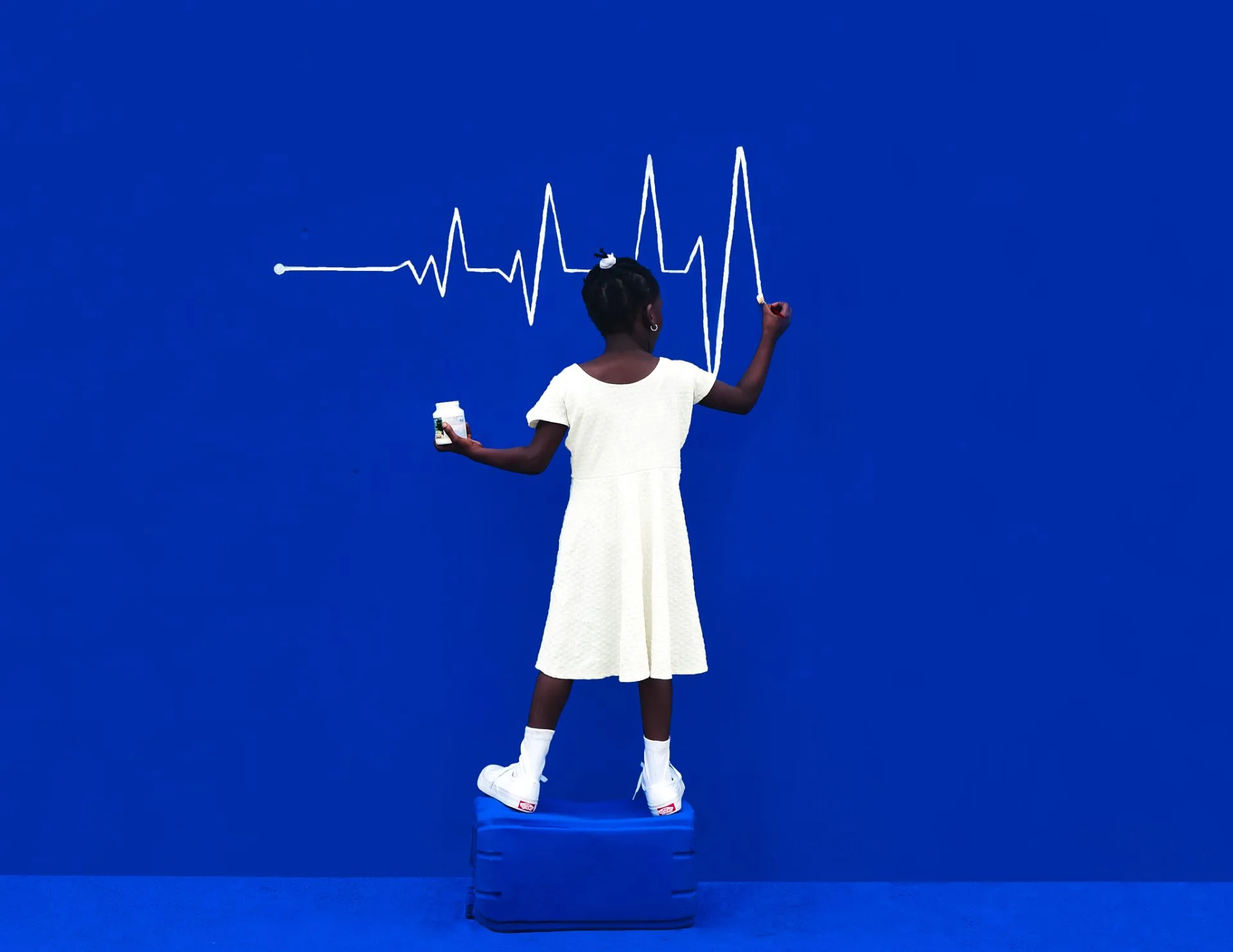 Une fillette en robe blanche, de dos sur un cube bleu, dessine à la craie un tracé de battements de cœur sur un mur bleu.