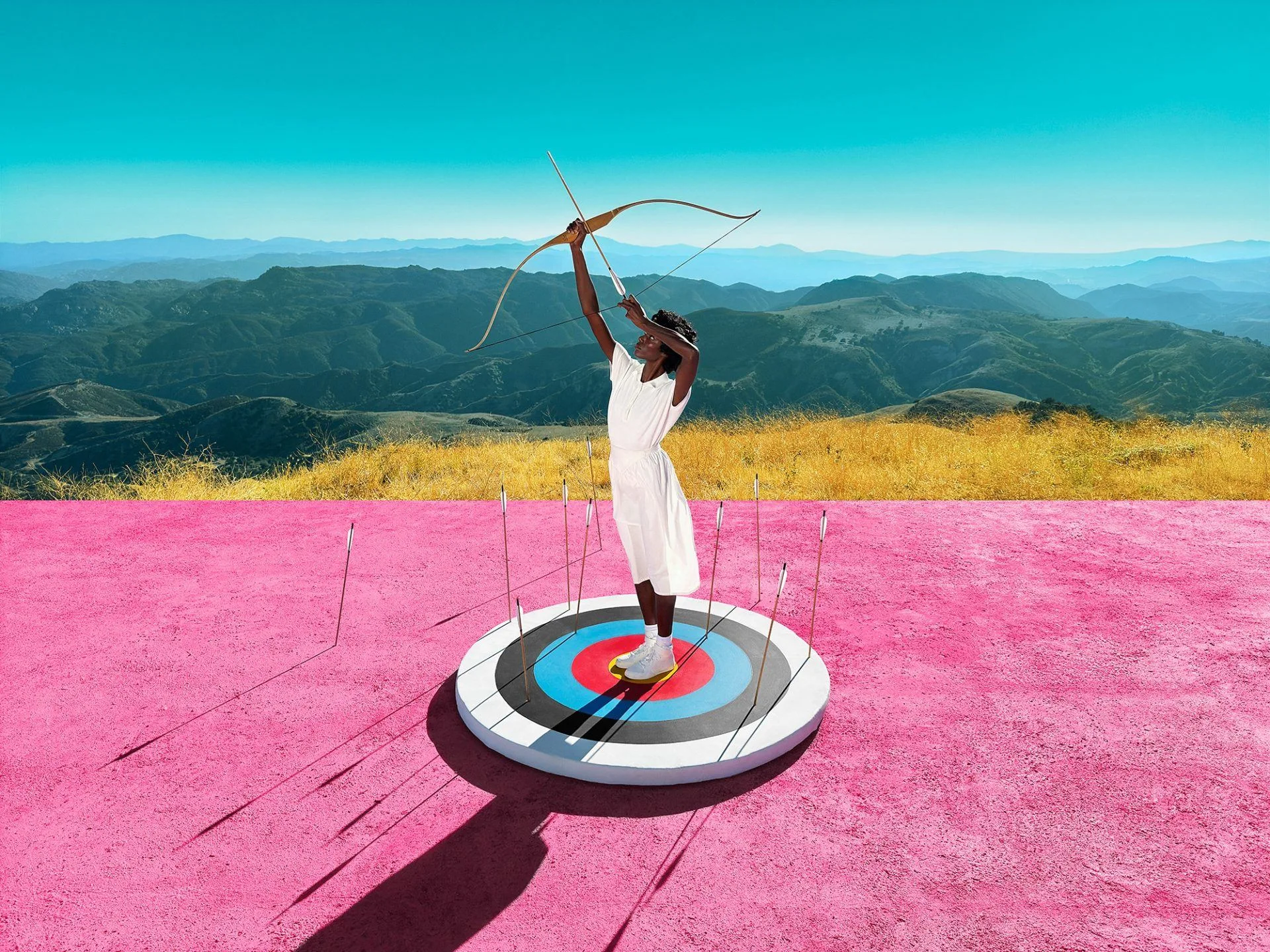 Une femme en robe blanche, debout sur une cible, tire une flèche vers le ciel sur un sol rose, devant des collines.