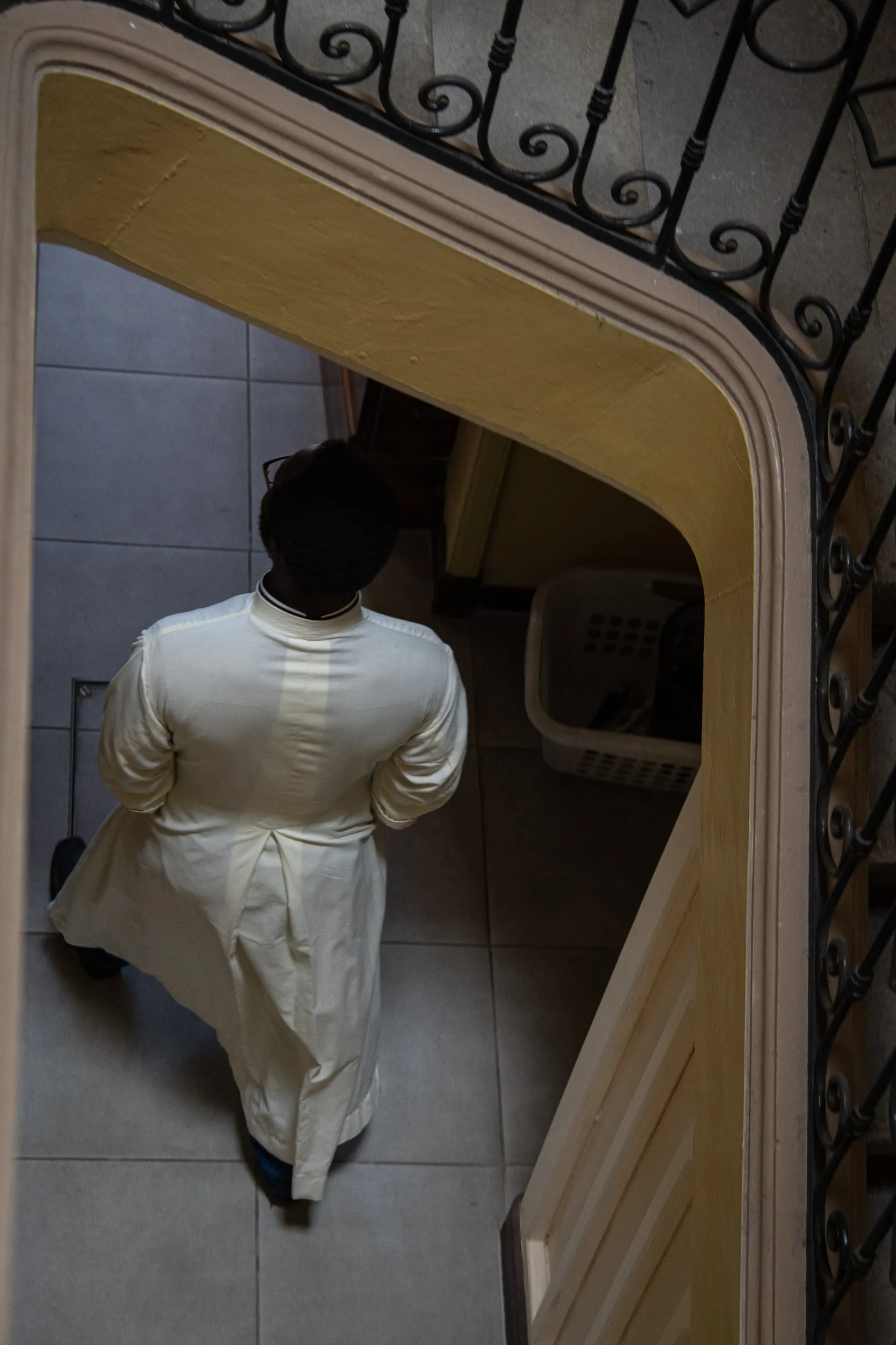 Vue plongeante dans une cage d'escalier à rampe de fer forgé : une personne en longue tenue blanche marche sur un carrelage.