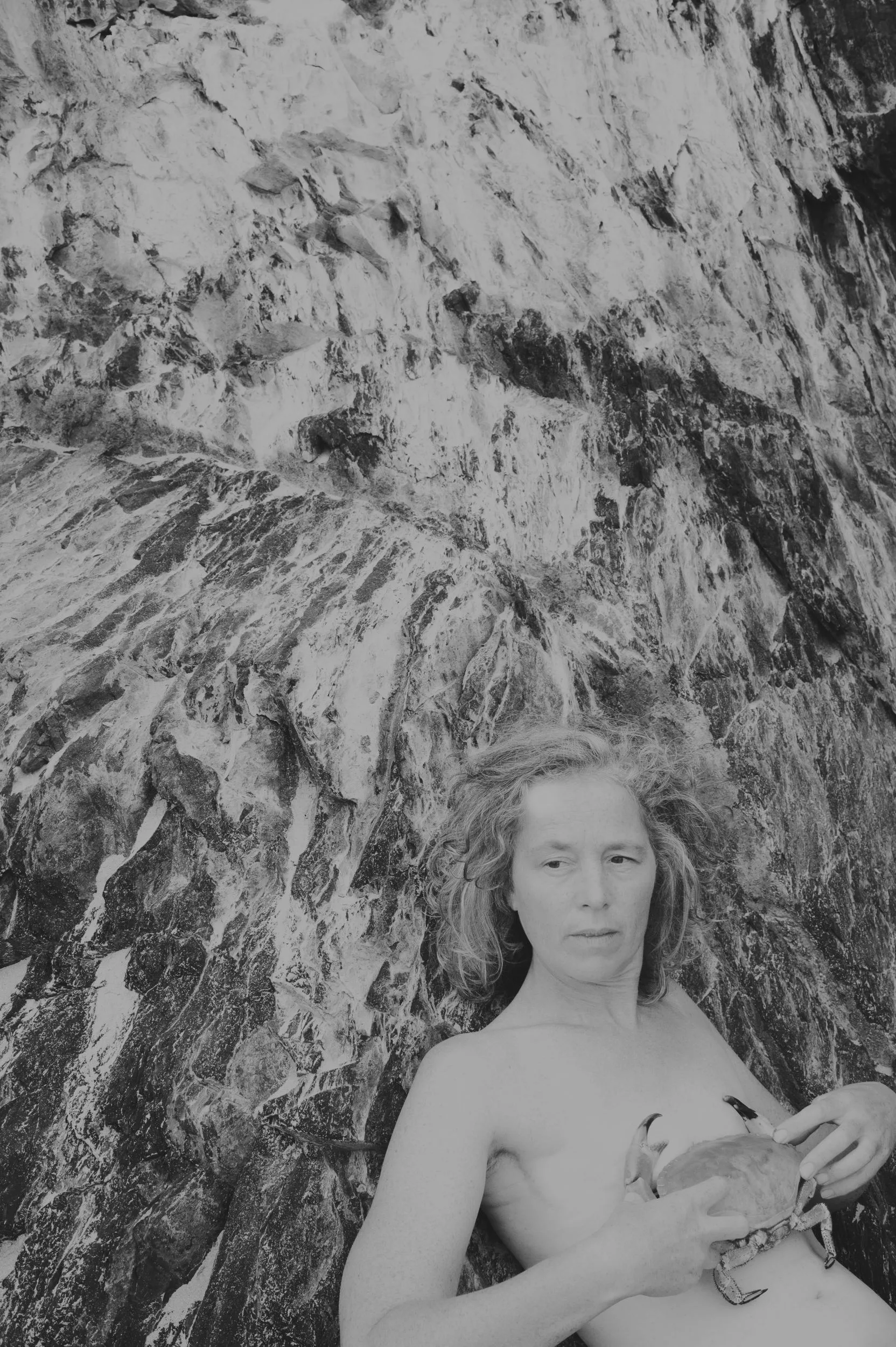 Photo noir et blanc : une femme torse nu, adossée à une paroi rocheuse, tient un crabe contre sa poitrine.