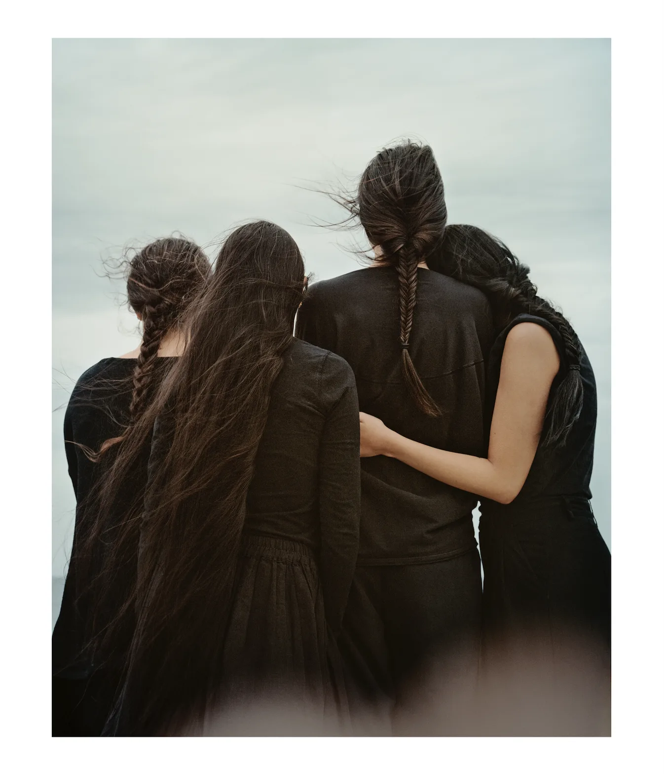 Quatre femmes vêtues de noir, vues de dos et enlacées, leurs longs cheveux tressés soulevés par le vent sous un ciel gris, par Hoda Afshar.