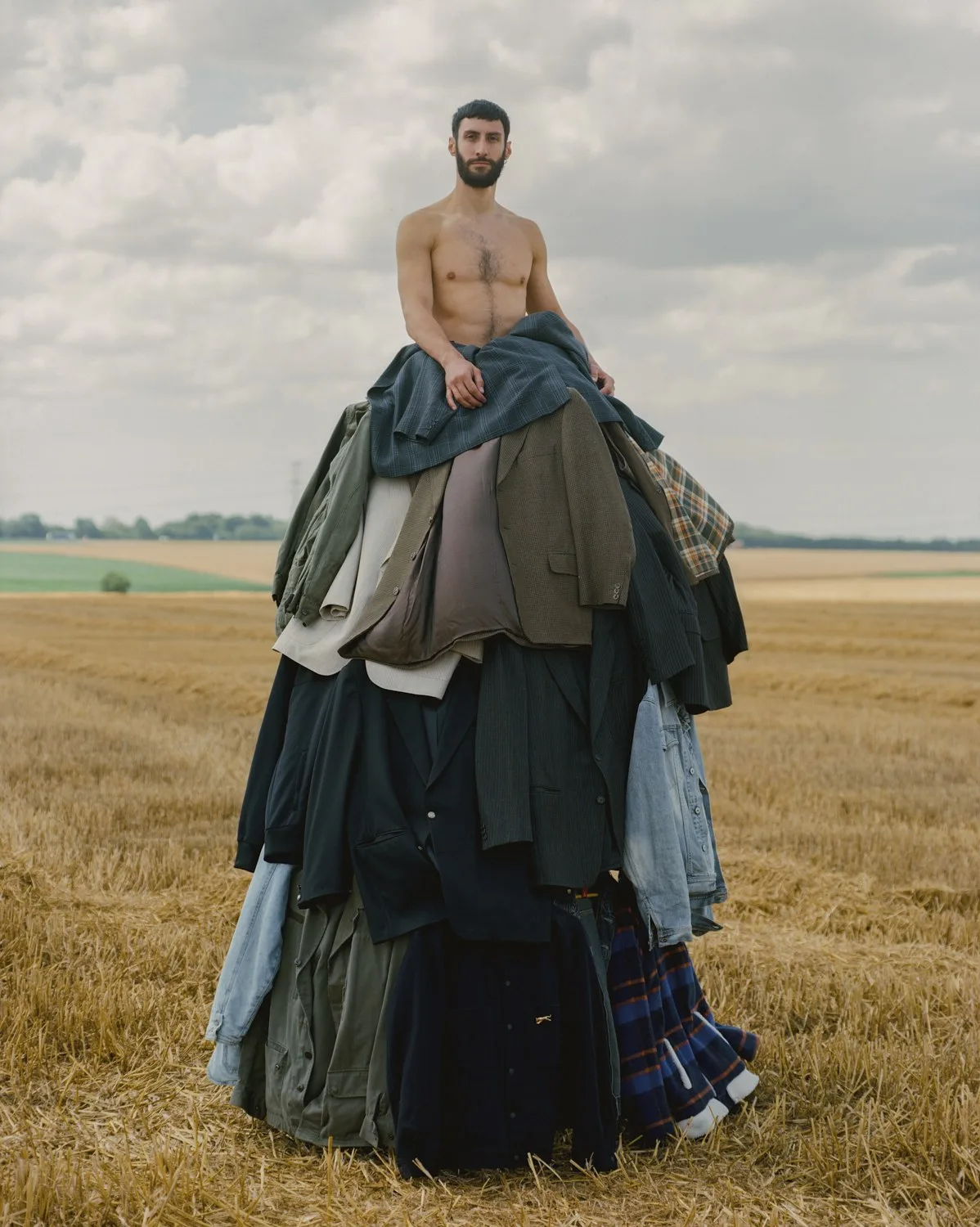 Un homme torse nu juché au sommet d'un haut tas de vestes et de manteaux usagés, au milieu d'un champ moissonné.