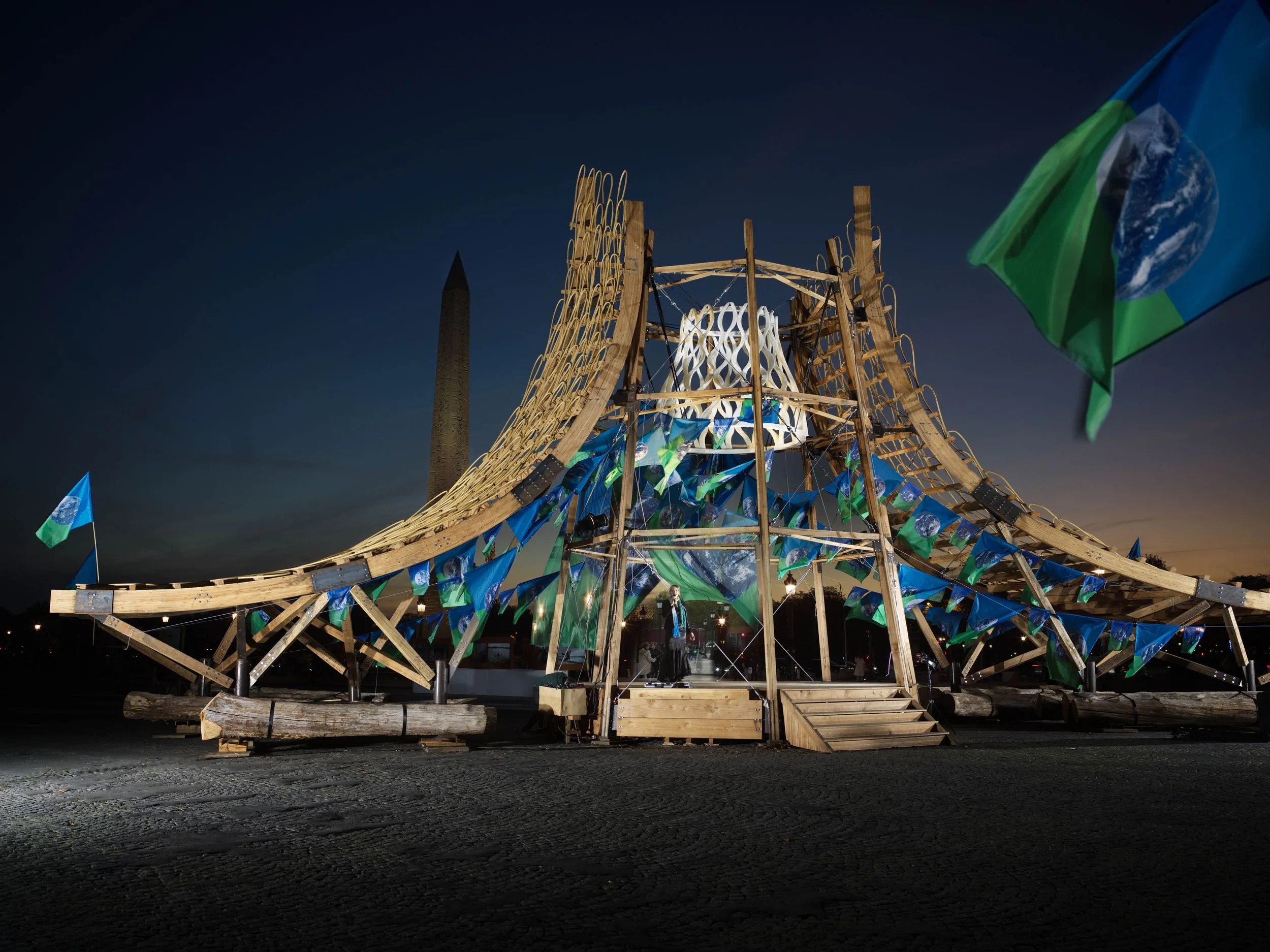 Le pavillon en bois « Concordia » d'Arthur Mamou-Mani la nuit, place de la Concorde, orné de drapeaux de la Terre, l'obélisque en arrière-plan.