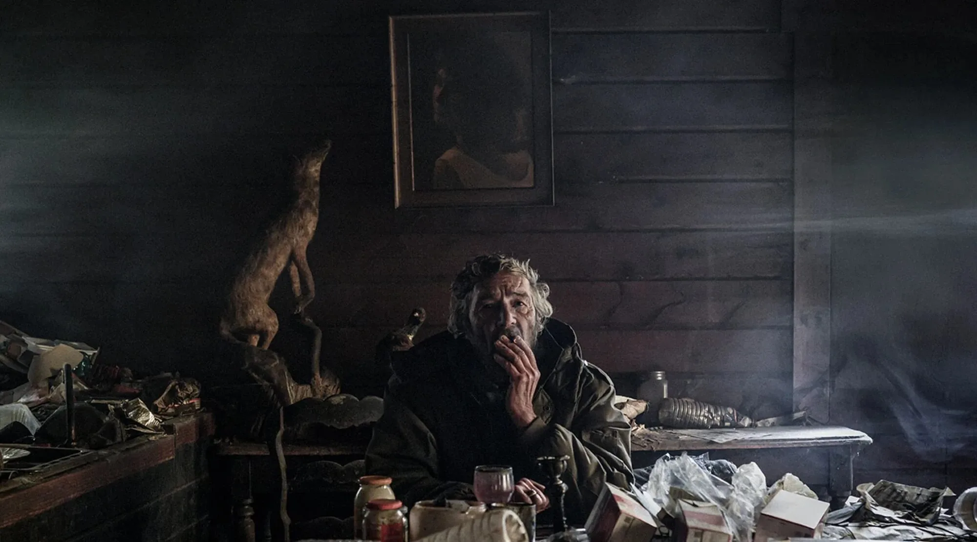 Un homme âgé fume, assis dans une pièce sombre aux murs de bois, encombrée de sacs et de cartons, sous un portrait encadré.